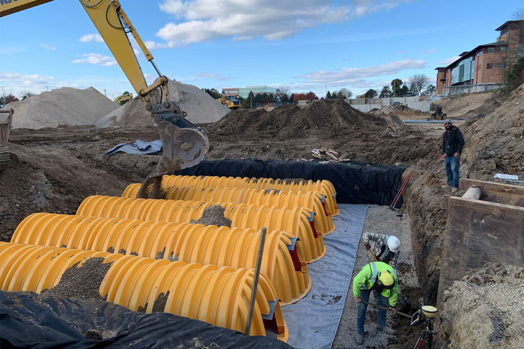 Workers installing underground stormwater chambers with an excavator backfilling gravel