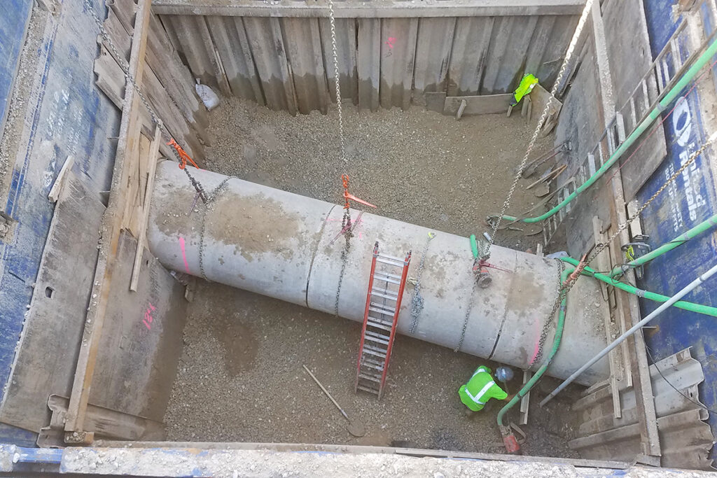 Concrete pipe being set into a trench with chains and workers below