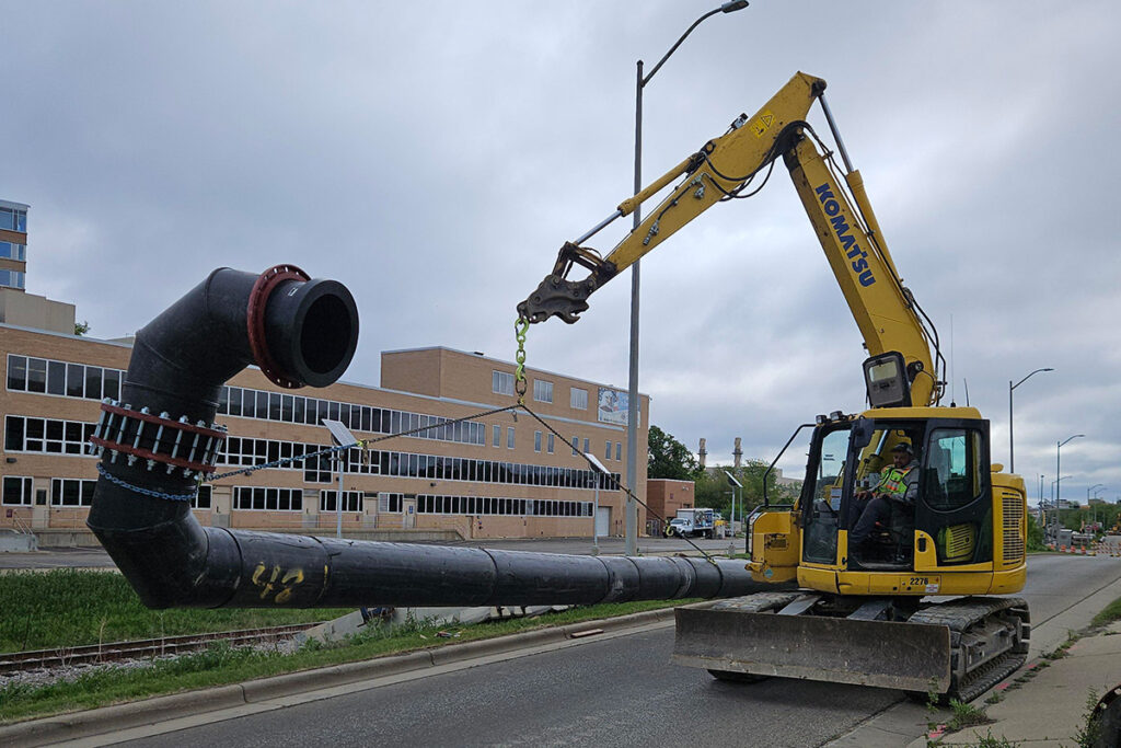 Excavator lifting and positioning a large industrial pipe over a roadway construction site